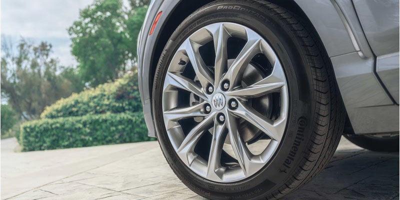 Buick car's front wheel with a Continental tire, parked on a paved driveway next to green bushes.