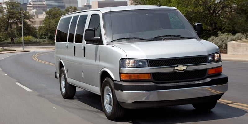 A silver Chevrolet Express passenger van drives along a curving road with a city skyline in the background.
