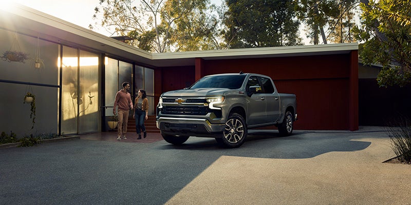 A light gray Chevrolet pickup truck parked in front of a modern home, with two people walking in the driveway