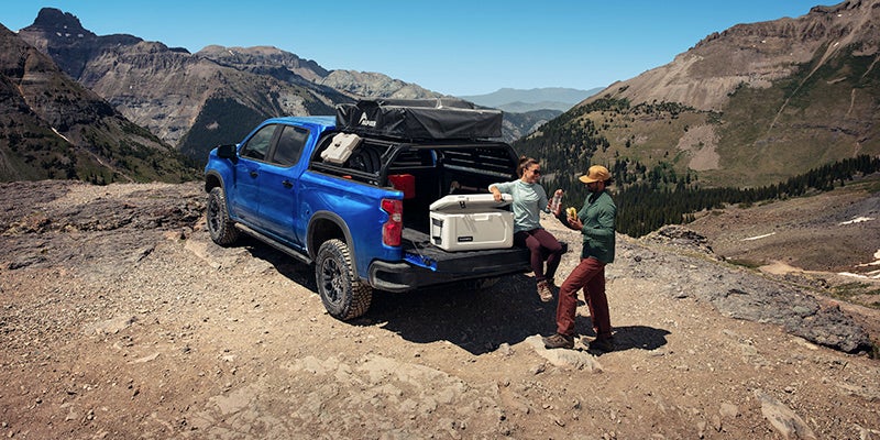 Blue Chevy pick-up truck parked on a mountain ledge with two people relaxing by the open tailgate in sunny weather.