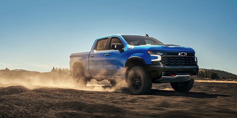 A blue Chevrolet Silverado ZR2 truck kicks up dust as it speeds across a dirt track under a clear sky.