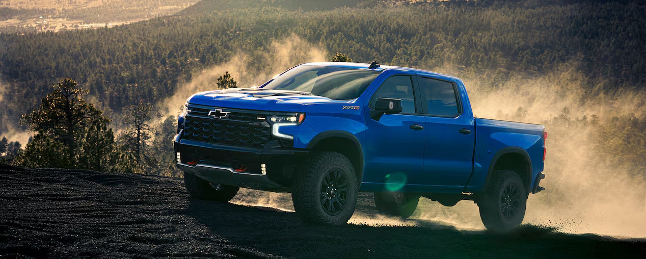 A blue Chevrolet Silverado ZR2 pickup truck drives on a dusty, rocky hill in a forest landscape.