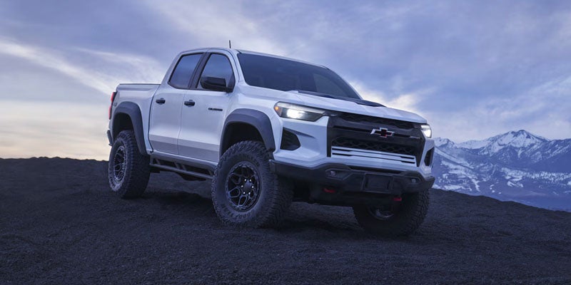 White 2023 Chevrolet Colorado ZR2 in a wide-angle shot, parked on dark gray terrain with mountains in the background.