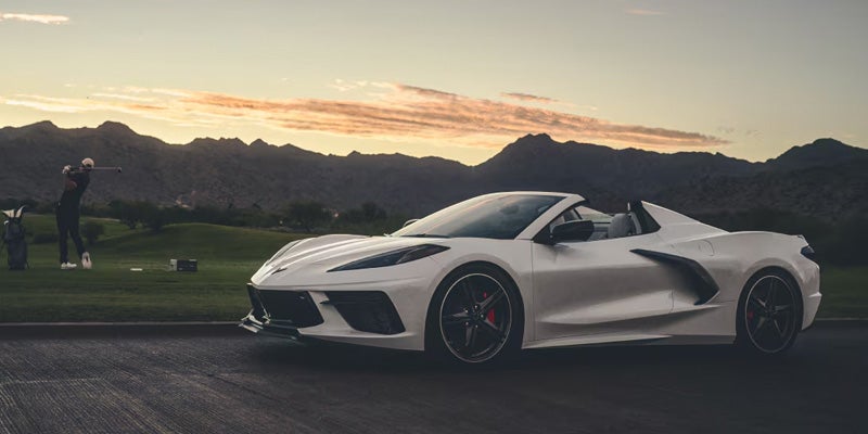Corvette convertible parked on a road with a golfer swinging on a course and mountains at sunset.
