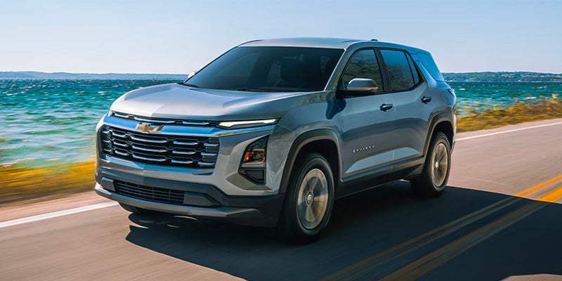 Front 3/4 view of a mid-size gray Chevrolet SUV driving on a coastal road with a blue, wavy ocean in the background.
