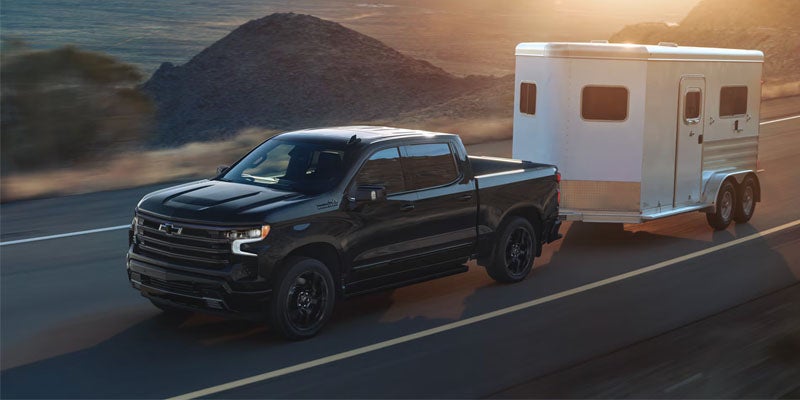 Black Chevy Silverado pickup truck pulling a silver horse trailer on a highway at sunset.