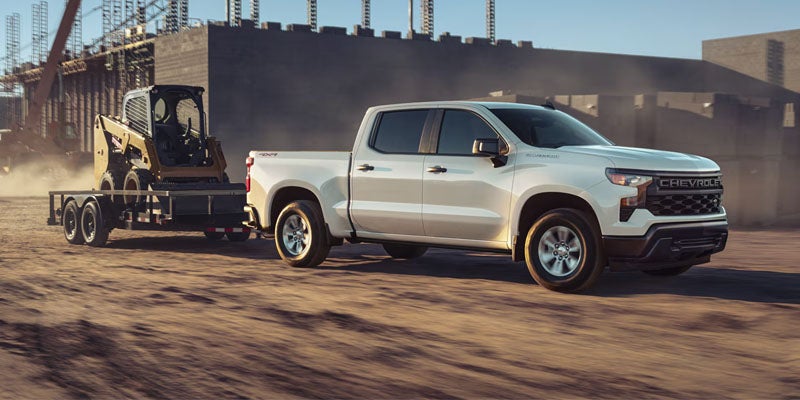White Chevrolet Silverado truck towing a trailer with a yellow skid steer in a dusty outdoor construction area.