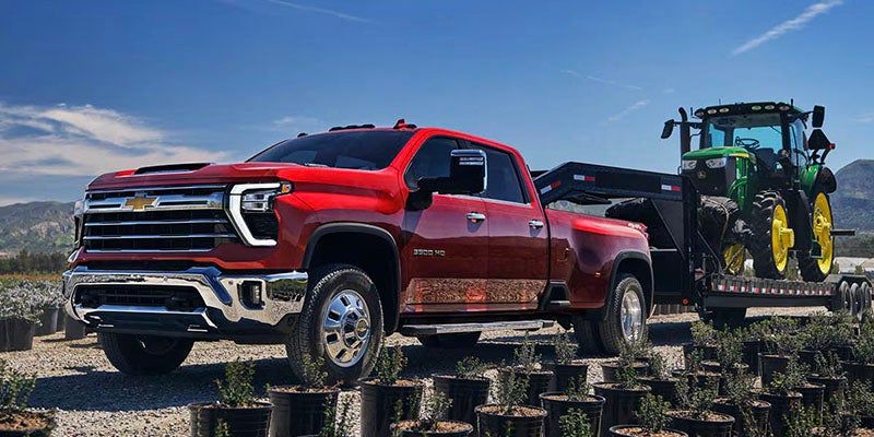 Red dually pickup truck towing a flatbed trailer with a green tractor in a field of potted plants.