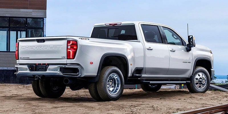 Rear and passenger-side view of a white Chevrolet Silverado Dually pickup truck on dirt next to a building.