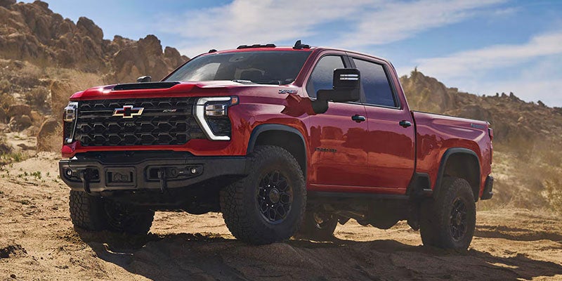 A dark red, lifted Chevy pickup truck with black wheels and a large front grille on a dusty, rocky dirt road.