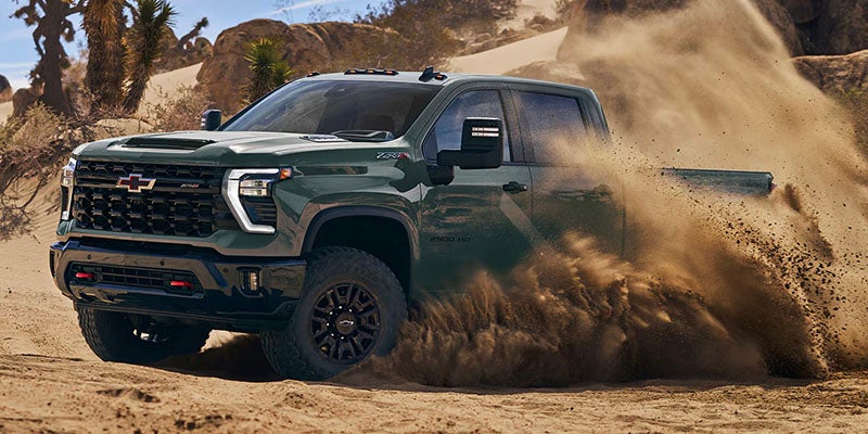 A dark green Chevrolet Silverado truck kicks up dirt while driving through a dry, desert landscape.