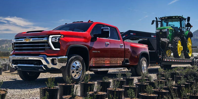 A red Chevrolet Silverado 3500HD pickup truck tows a trailer with a green John Deere tractor in a field of plants.