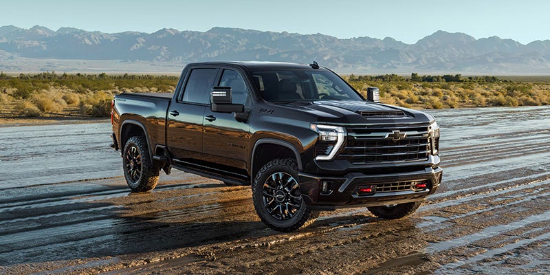 A black Chevrolet Silverado HD pickup truck with a large grill and off-road tires, parked on a muddy road in the desert.