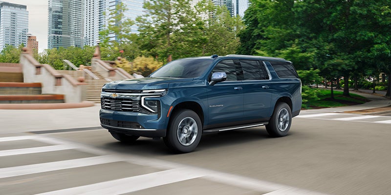 A dark blue Chevrolet Suburban SUV drives left across a city street, with buildings and trees in the background.