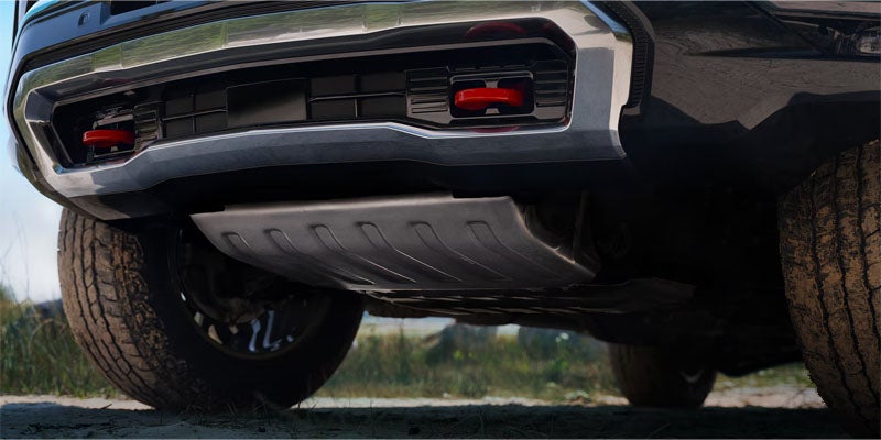 Underside of a black pickup truck with two red tow hooks, a silver skid plate, and knobby tires on dirt.
