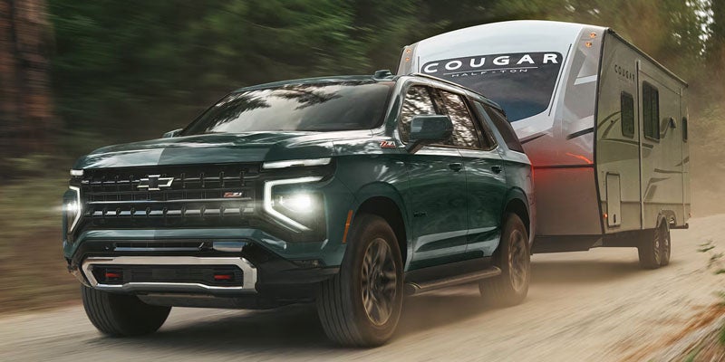 Green Chevy Tahoe SUV towing a trailer on a dirt road surrounded by trees.
