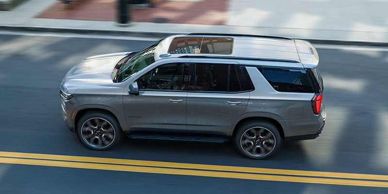 Silver Chevrolet Tahoe SUV driving on a paved road viewed from above with motion blur.
