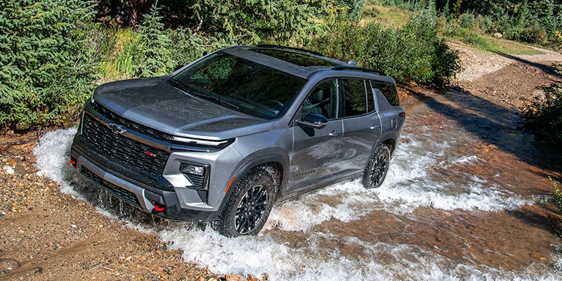 A gray SUV with a black grill and red accents drives through a shallow stream in a forest.