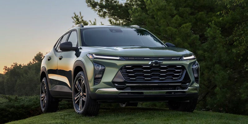 A green Chevrolet Trax Activ 2024 parked on a grassy hill with green trees and sunset in the background.