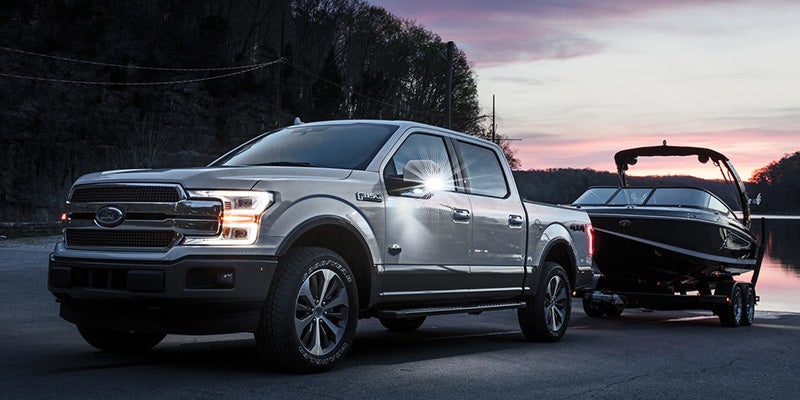 A silver Ford F-150 pickup truck tows a boat on a trailer down a ramp towards a lake at sunset.
