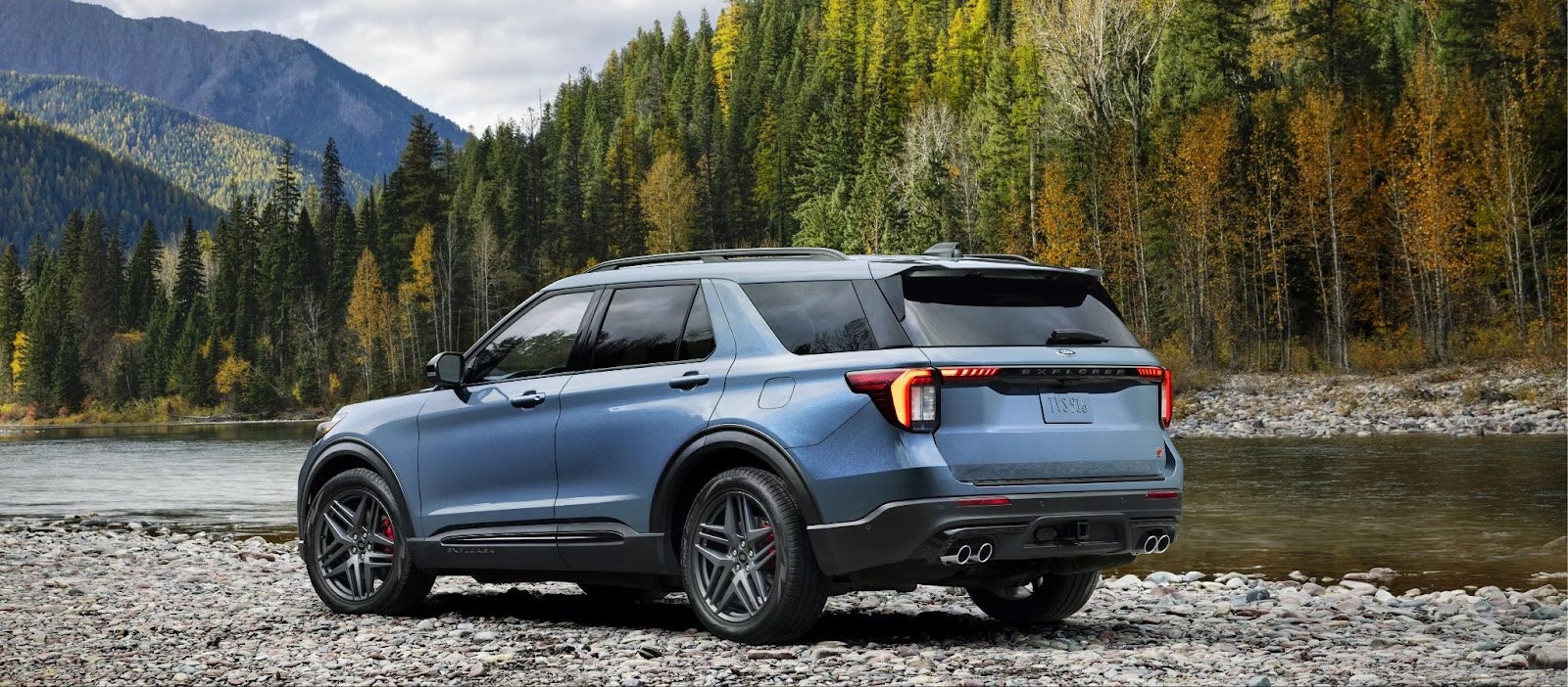 Rear-side view of a blue 2024 Ford Explorer ST on a rocky riverbank, mountains and forest in background.
