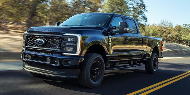 A dark blue Ford F-Series Super Duty pickup truck driving on a winding road with trees in the background.