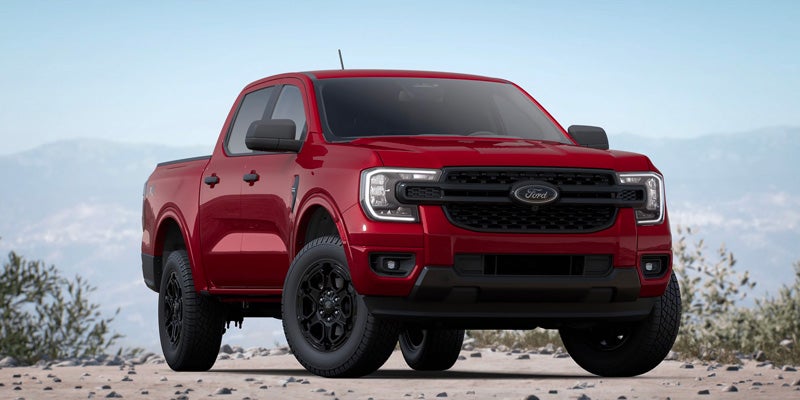 Front-facing view of a cherry-red Ford Ranger pickup truck with black wheels on a dirt road, mountains in background.