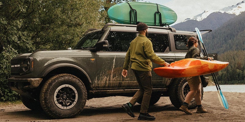 A person and child unload a kayak and surfboard from a parked SUV near a lake and mountains.