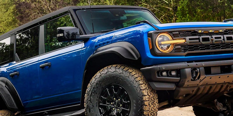 A blue Ford Bronco drives over a dirt pile, highlighting its off-road capabilities and bold front grille.