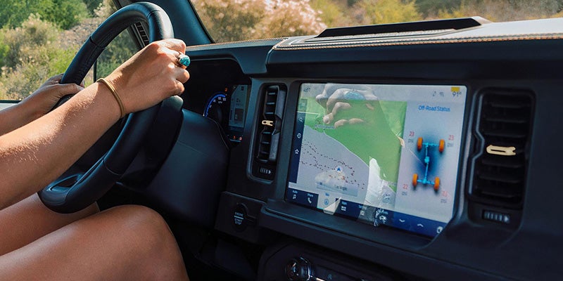 A person's hand rests on the steering wheel of a car, with a navigation map displayed on the dashboard screen.