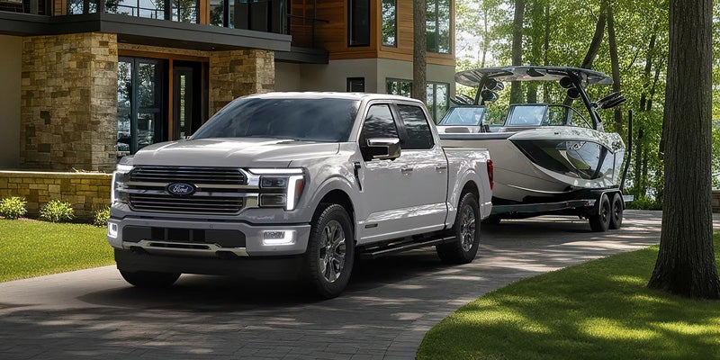 Ford F-150 truck with white LED headlights, towing a boat on a trailer, parked on a paved driveway in front of a modern stone and wood house.