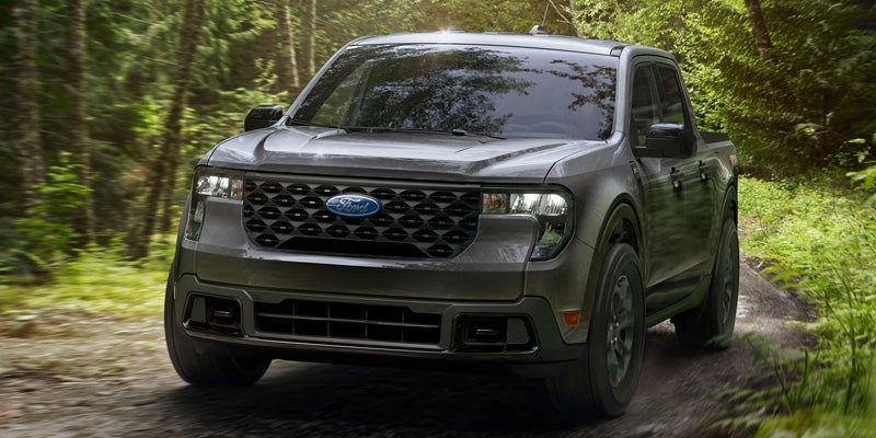 Dark gray Ford Maverick pickup truck driving on a dirt road through a sun-dappled forest.