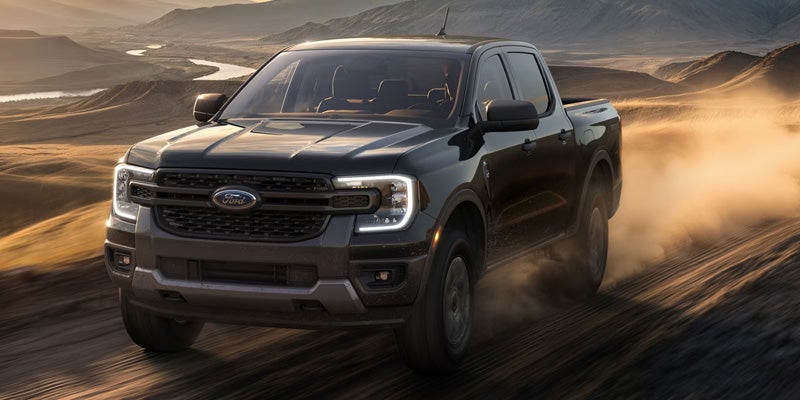 Black Ford Ranger pickup truck driving on a dirt road, kicking up dust, with mountains and a river in the background.