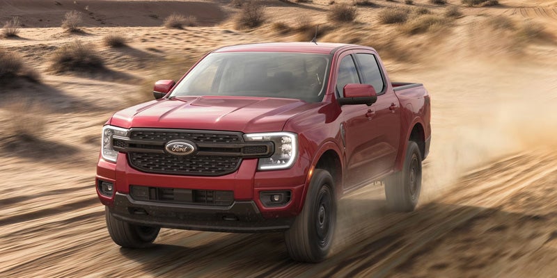 A red Ford Ranger pickup truck kicks up sand as it drives over desert dunes.