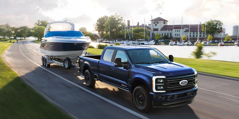 A dark blue Ford F-Series pickup truck tows a boat on a trailer down a road beside a marina filled with sailboats.