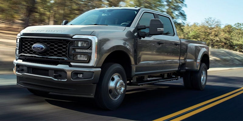 A gray Ford F-Series Super Duty truck driving down a sunny road with trees in the background.