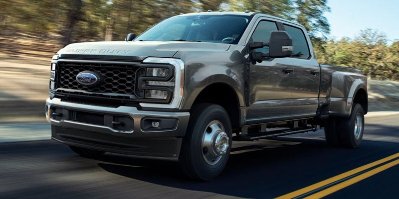 A dark gray Ford F-350 Super Duty dually pickup truck drives on a two-lane road lined with trees.