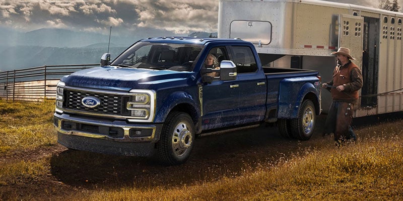 A blue Ford Super Duty truck with a woman driving pulls a livestock trailer, as a cowboy stands outside, on a rural road.