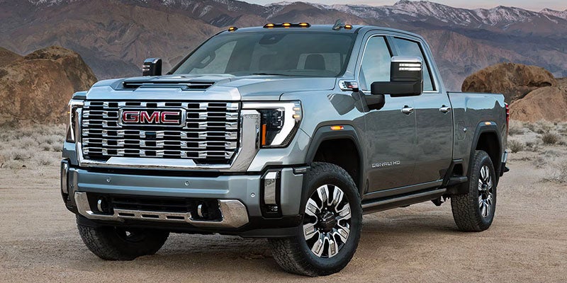 A silver GMC Denali HD pickup truck with chrome grille and accents, parked on a dirt road in a desert landscape.