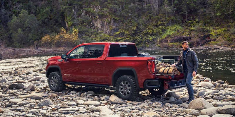 A red pickup truck parked on rocky terrain beside a river, with a man standing near its tailgate.