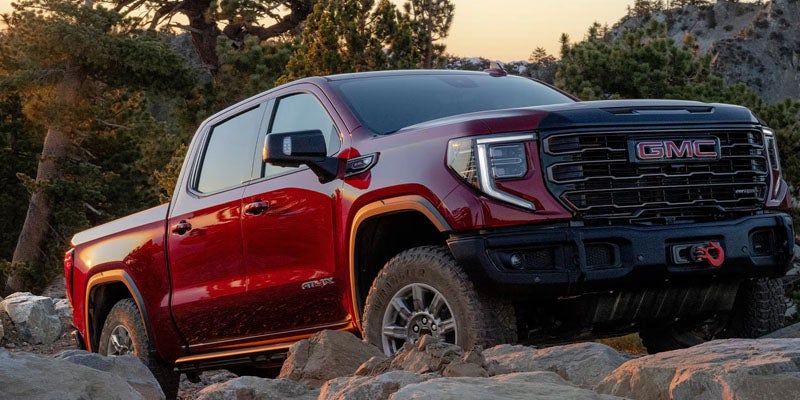 GMC pickup truck with black trim drives over rocks in a wooded, mountainous area at golden hour.