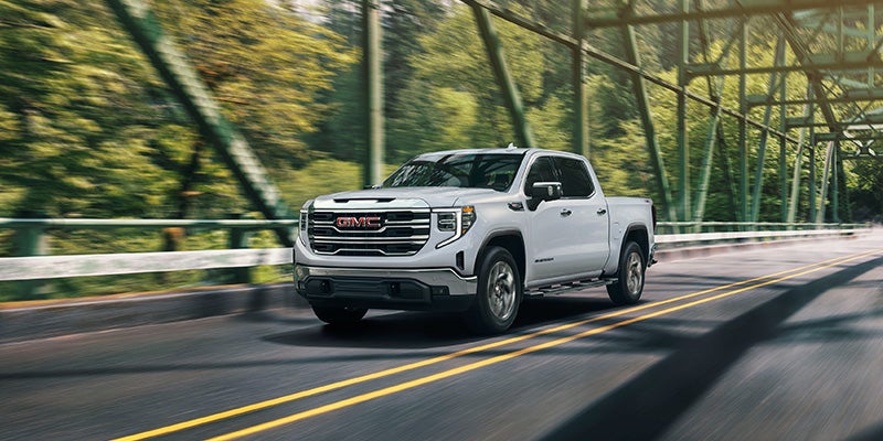 A silver GMC Sierra pickup truck drives across a metal bridge surrounded by green trees.