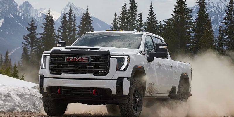 A white GMC Sierra AT4X truck drives on a dirt road, kicking up dust, with snowy mountains in the background.