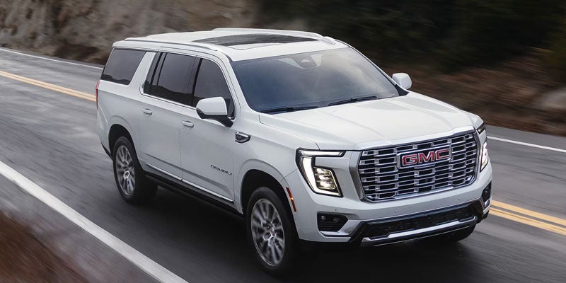 Front quarter view of a white GMC Yukon Denali SUV driving on a wet road, with mountains in the background.