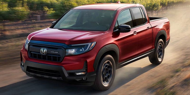 A red Honda Ridgeline pickup truck driving on a dirt road, kicked-up dust visible behind its black wheels.