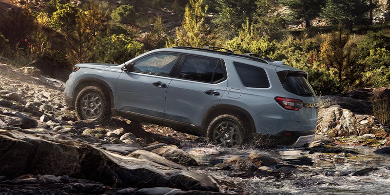Steel blue SUV with black roof rails on rock-strewn terrain, crossing a shallow stream with forest in background.