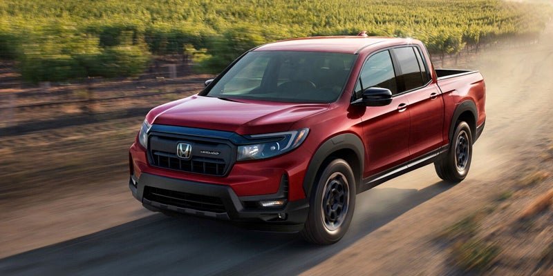A red Honda Ridgeline pickup truck driving fast on a dirt road, with green fields in the background.