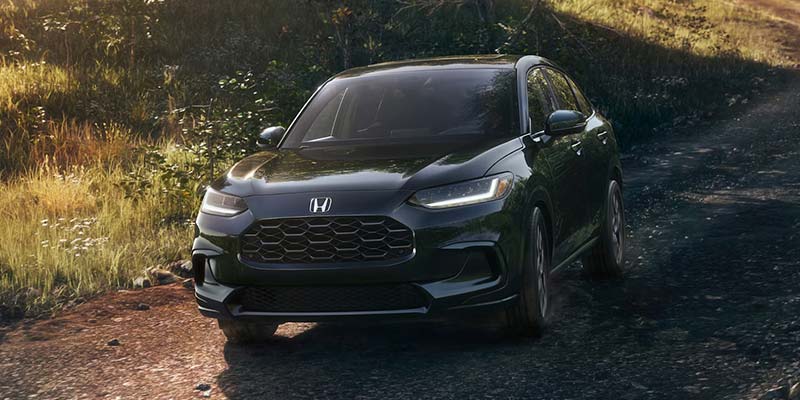 Front view of a dark grey Honda HR-V with LED daytime running lights, parked on a dirt road surrounded by greenery.