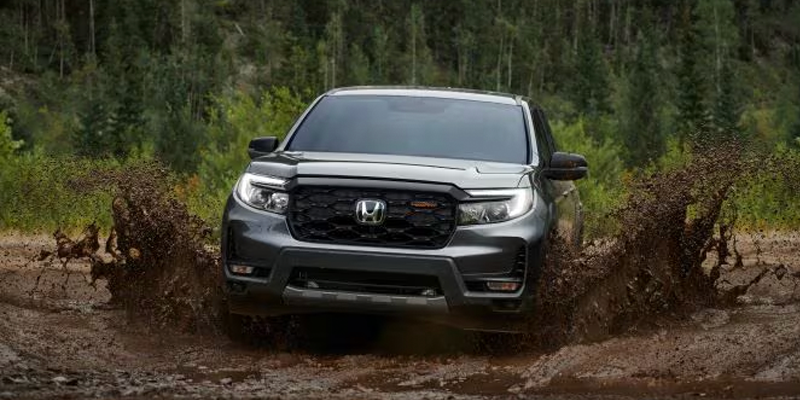 Dark gray SUV driving through a mud puddle, splashing mud high into the air, with a forest in the background.