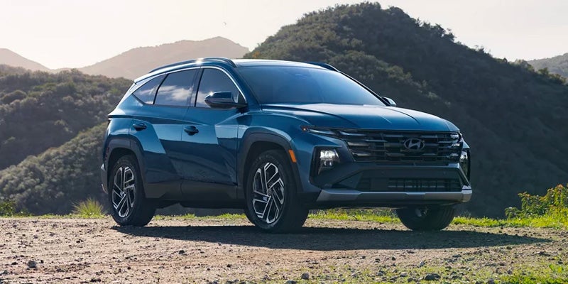 A dark teal Hyundai Tucson SUV, facing slightly right, parked on a dirt path with treed mountains in the background.